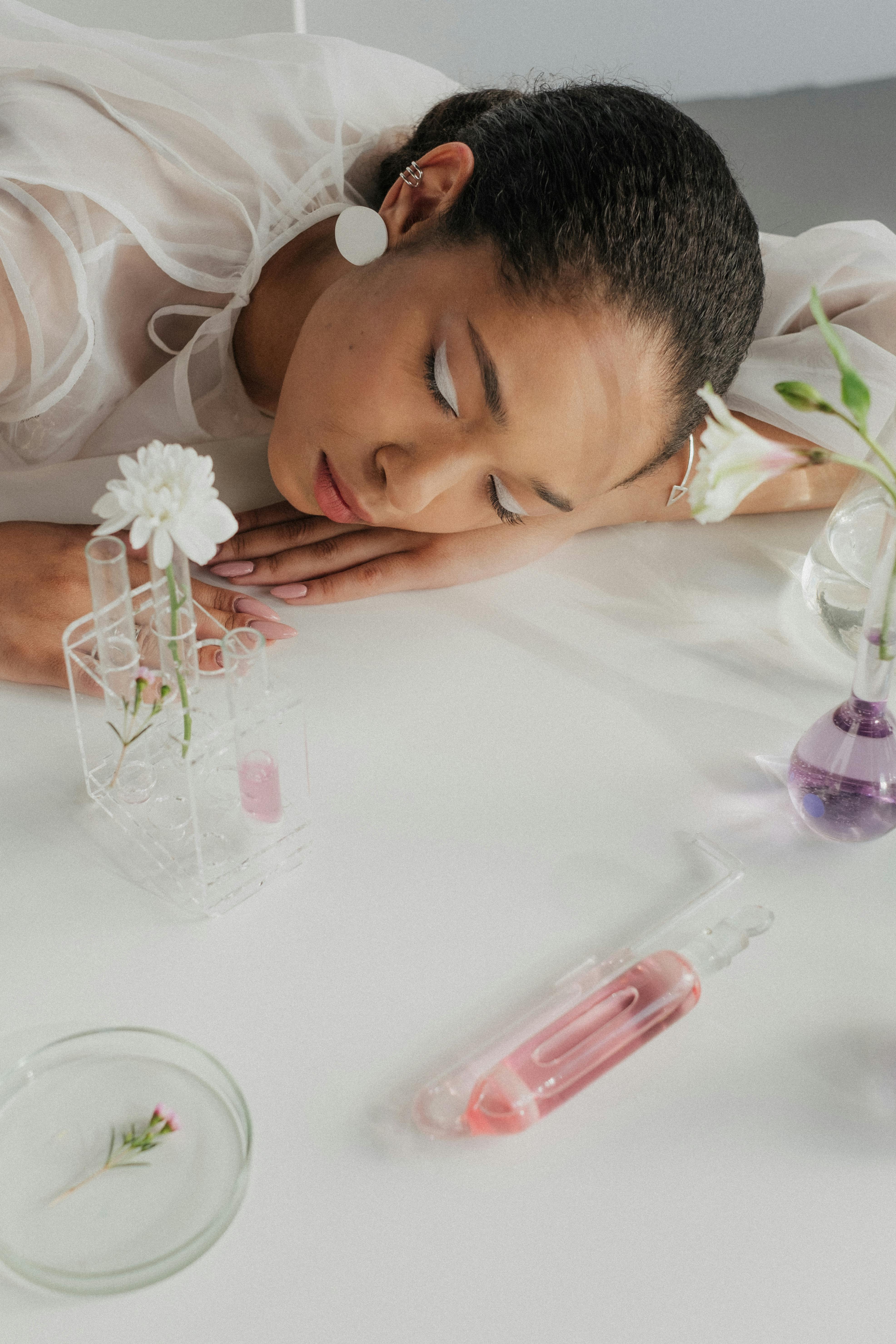 A woman rests on a table surrounded by glass flasks and flowers in a laboratory setting